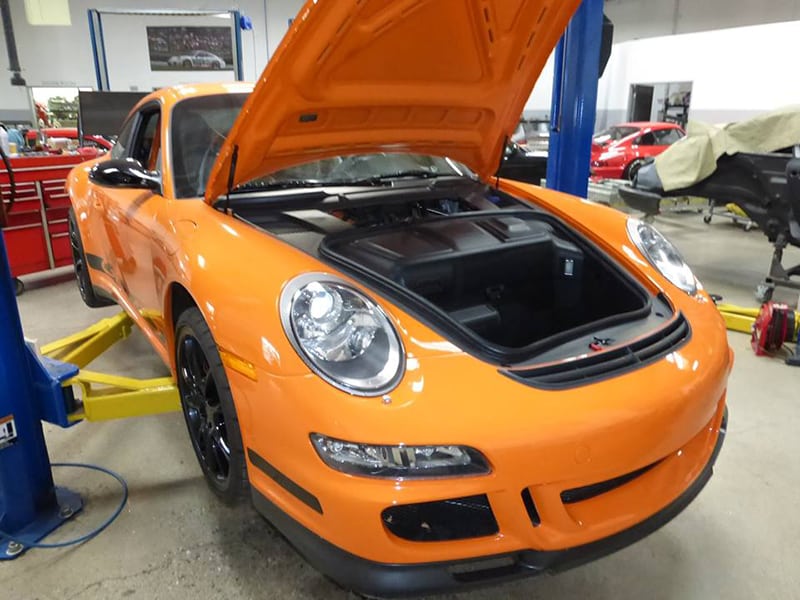 Orange Porsche 911 GT3 RS with frunk open on a garage lift inside a shop.