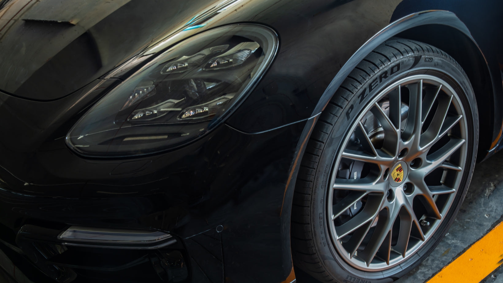 Close-up of a black Porsche Panamera headlight and wheel with a visible Porsche crest logo on wheel center cap.