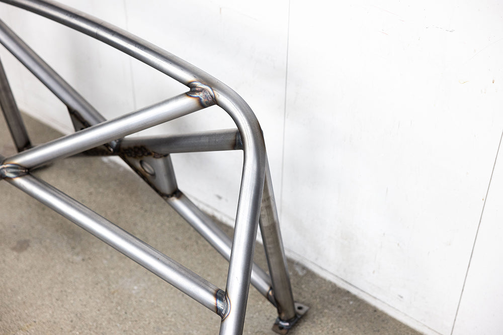 Close up of 911 Design roll cage in raw metal on shop floor against white wall.
