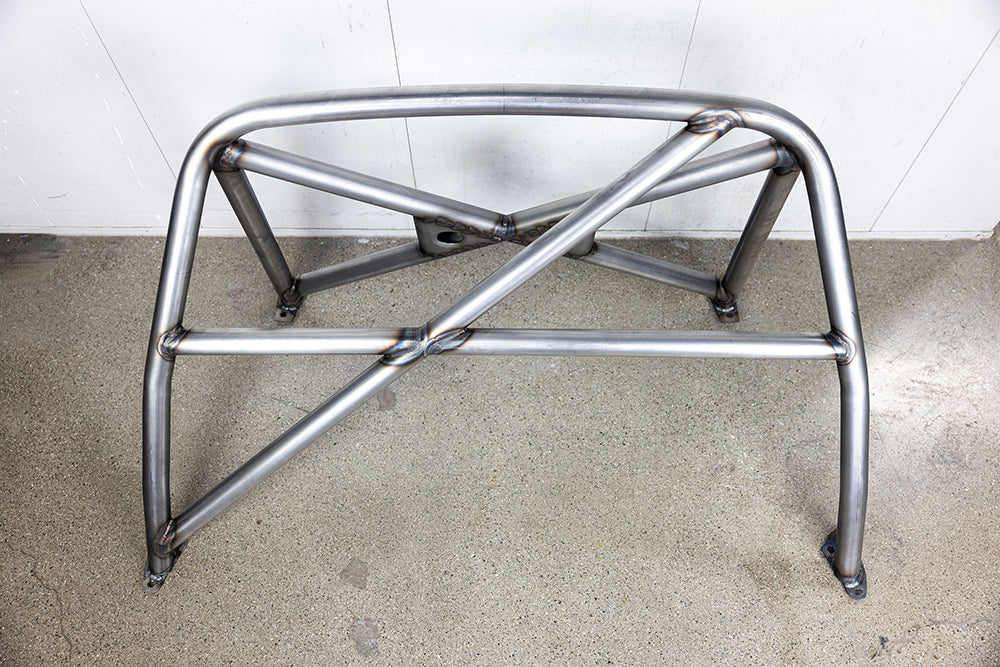 911 Design roll cage in raw metal on shop floor against white wall.