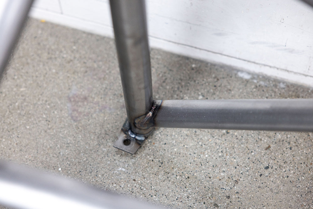Close up of 911 Design roll cage in raw metal on shop floor against white wall.
