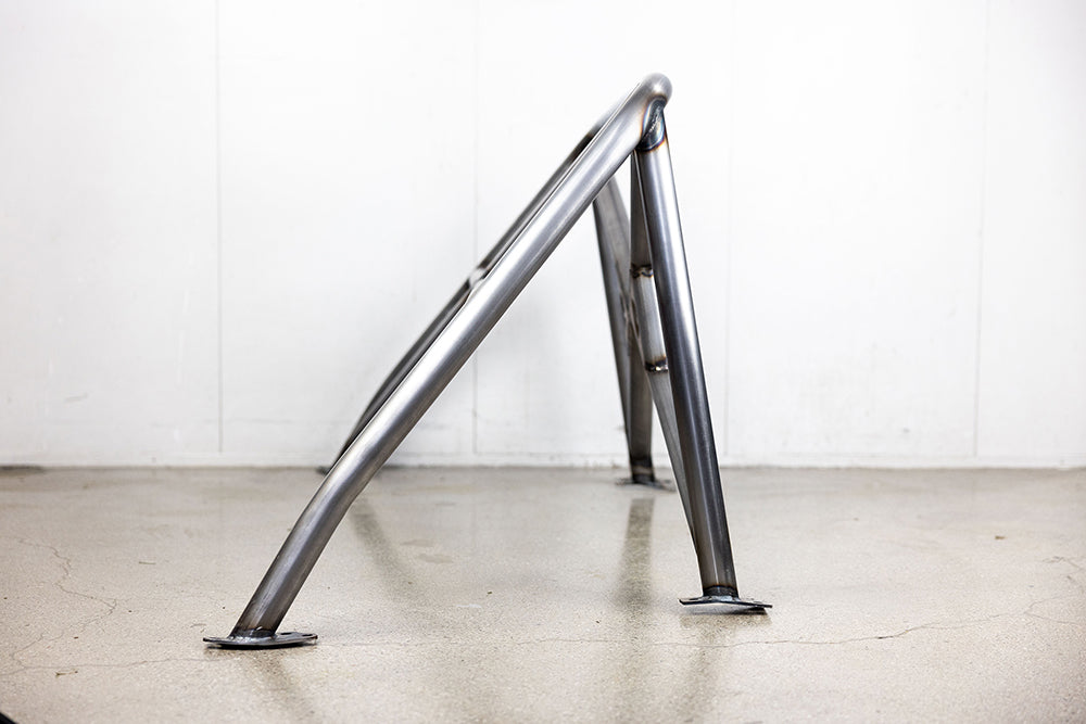 Side view of 911 Design roll cage in raw metal on shop floor against white wall.