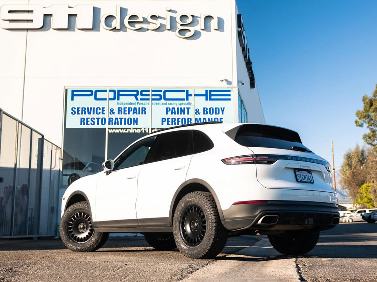 White Porsche Cayenne SUV parked in front of a 911 Design service and repair building.