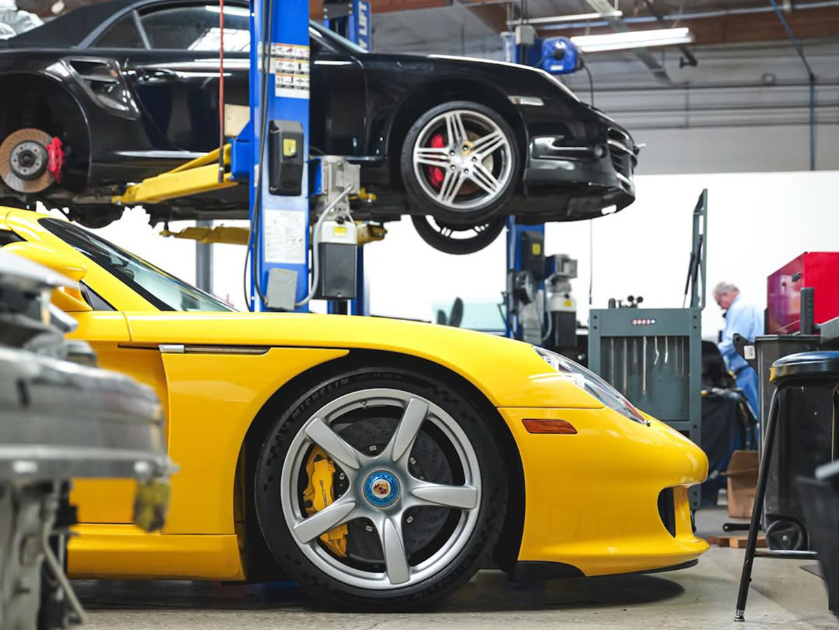 Yellow Porsche Carrera GT side profile inside garage with black 911 Turbo Cabriolet on lift in background.