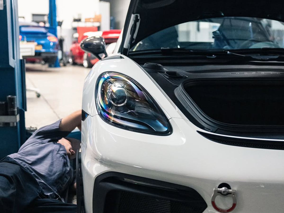 Close-up of a Porsche Cayman front headlight with a technician working on it inside a garage.