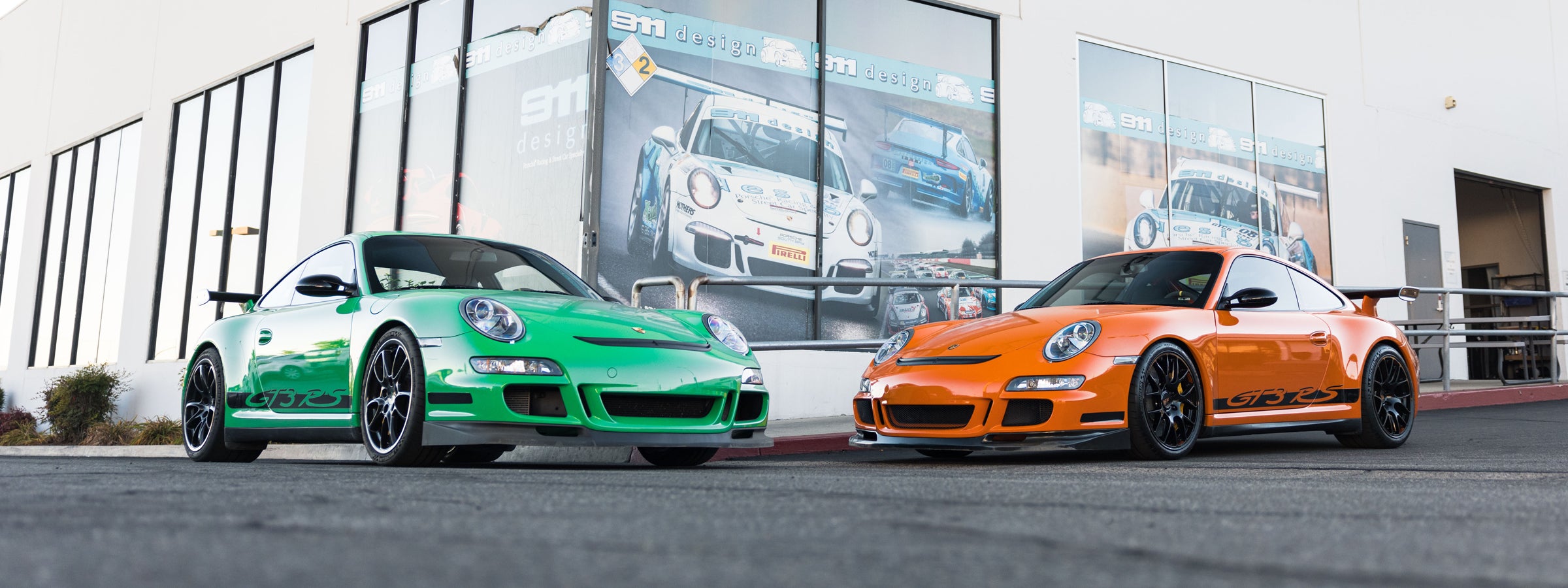 Two 997 GT3s, one green and one orange, parked in front of 911 design building.